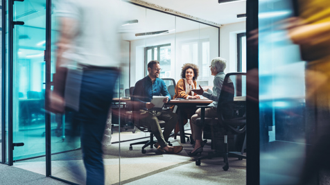 Three colleagues engaged in a discussion inside a modern glass-walled office meeting room.
