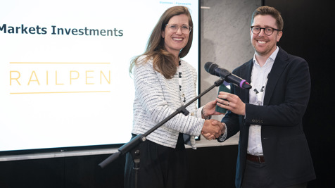 A man and a woman in smart attire posing for a photograph in front of the screen. The screen features Railpen logo and says Markets Investments