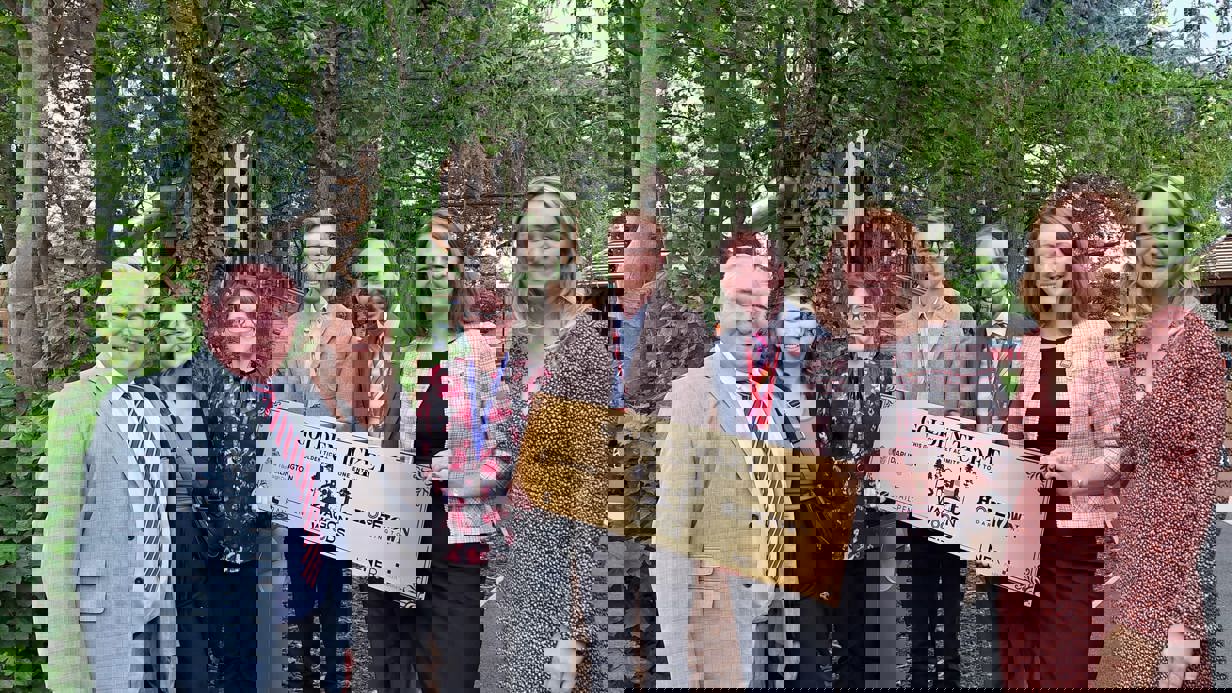  group of people standing outdoors holding a large “Golden Ticket” plaque, gathered together in a garden setting with trees and wooden play structures in the background.