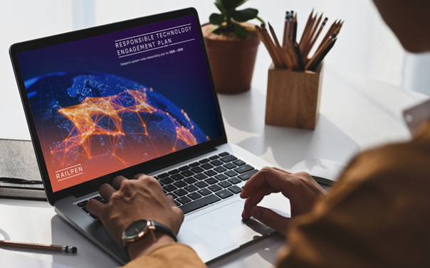 A person sits at a desk using a laptop displaying the cover of Railpen’s Responsible Technology Engagement Plan. The screen shows a digital globe with interconnected orange and yellow network lines, the Railpen logo, and the report title. The workspace includes a plant, a pencil holder with pencils, and a bright, modern background.