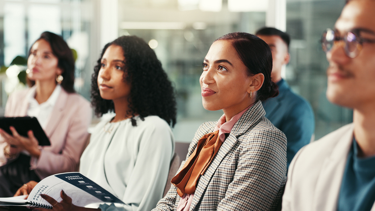 Colleagues seated in a row holding workbooks look up to a stage