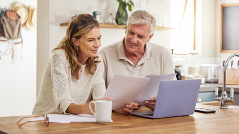 Man and woman sit in a kitchen reading papers and laptop screen