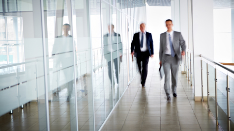 Two businessmen walking through a modern office corridor with glass walls and tiled floors.
