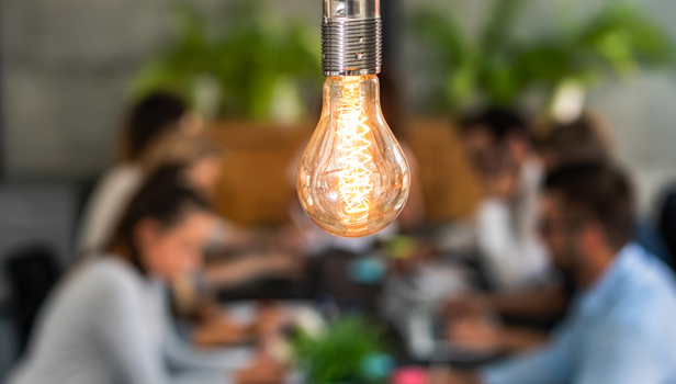 a flashing lightbulb at the forefront and people working in the office at the background