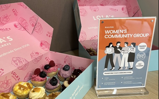 Open pastel‑blue boxes of assorted mini cupcakes are displayed on a wooden table, with colourful toppings like berries and swirls of frosting. Next to the boxes stands an upright sign featuring illustrated figures and the heading “Railpen Women’s Community Group,” promoting group activities and training opportunities.
