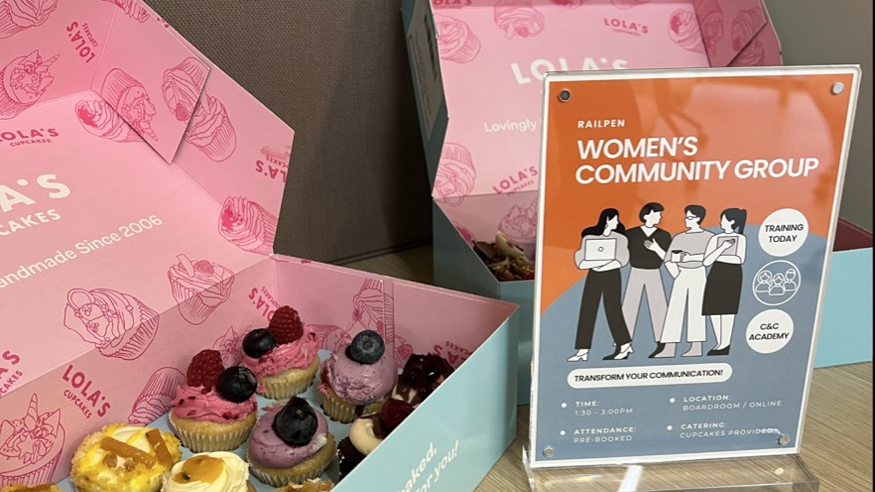 Open pastel‑blue boxes of assorted mini cupcakes are displayed on a wooden table, with colourful toppings like berries and swirls of frosting. Next to the boxes stands an upright sign featuring illustrated figures and the heading “Railpen Women’s Community Group,” promoting group activities and training opportunities.